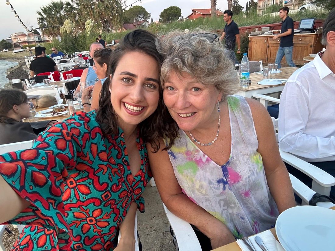 Selin and Luke&rsquo;s mum at a long table by the sea, heads together, both laughing